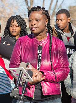 A woman with dreaded black hair and a purple leather jacket speaks toward TV cameras.