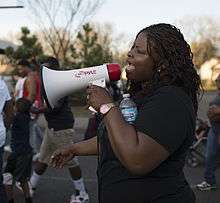 A woman with black hair in a black T-shirt speaks through a megaphone as she marches.