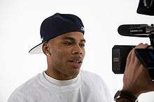 A man in front of a handheld video camera, wearing a white T-shirt and his cap backwards