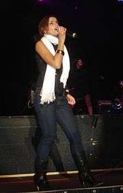 Nelly Furtado wearing a black singlet, a white scarf, blue jeans and black boots, singing into a microphone