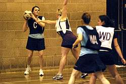 Female netball player in dark bib holding the ball with her hands positioned to make a two handed pass. She is being guarded by another female player in a white bib.