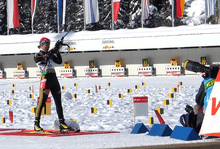 A woman in predominantly black winter sportswear, looking concentrated, holds a rifle which points into the sky. She stands on a red mat and a photographer with a large object lens kneels on the right hand side. Parts of a shooting range covered in snow can be seen in the background.