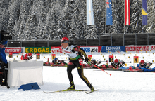 A woman on cross country skies wearing a red cap, a white jersey with the number 15 and black trousers skies away from a shooting range covered in snow. Half a dozen people in the background are shooting while lying on the ground.