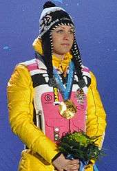 A woman, wearing a predominately yellow and pink jacket and a black cap, stands in front of a blue background, looking to the right. She hold flowers in her hands and has a gold medal around her neck.