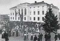 People gathered in front of the new city hall, which is white with two stories and stately features