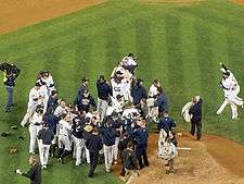 Many men in white baseball uniforms and blue caps, some wearing blue jackets, stand near a dirt mound. Some are hugging each other.