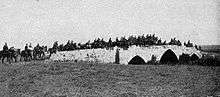 Photo shows brigade crossing the three arched stone bridge (two smaller arches on either side of a central larger arch) with the road grading up to the center and down on the ends.