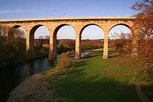 A large bridge crossing a river, and the rivers floodplain beside it. The bridge has 7 visible arches, and is taller than the mature deciduous trees surrounding it.