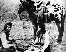 In the foreground, two Native American men wearing cowboy attire sit crosslegged on the ground. In the background, a dark colored horse with a white and black spotted rump stands saddled and bridled.