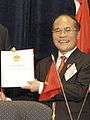 a smiling nearly bald man, wearing glasses, a suit and a red tie while holding a piece of paper