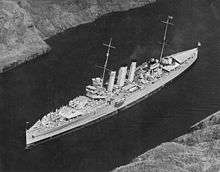 Aerial photograph of a cruiser-size warship sailing slowly through a narrow body of water