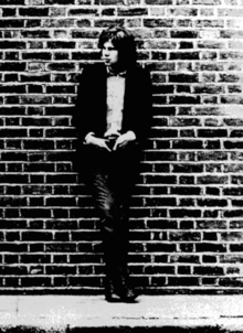 A black-and-white photograph of a man leaning against a brick wall, with his hands resting on his waist.