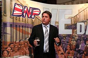 man in a suit holding a pint of beer standing in front of sign saying 'BNP VE Day'