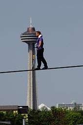 A man with a purple shit and black pants tightrope walks with a large tower in the background