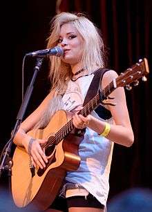 Nina Nesbitt toting an acoustic guitar before a microphone on a stand.