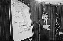 A room with a curtain and an American flag in the background. A man in a suit points to Cambodia on a large standing map of Southeast Asia.
