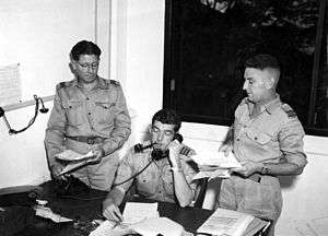 Three air force officers in light-coloured summer uniforms, one seated at a desk and the others standing beside him