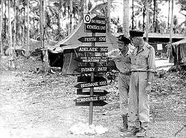 Two men in light-coloured uniforms with a sign reading "Perth 5290, Adelaide 3507, Young & Jacksons Pub Melbourne 2990, Sydney 2472, Brisbane 1955, Townsville 1237"