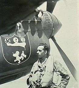 Moustachioed man in flying gear standing beneath the nose of a single-engined fighter plane, which pictures a dog and a swastika, among other items