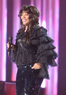 With long brown hair, a woman smiles and holds a microphone, wearing a black outfit.