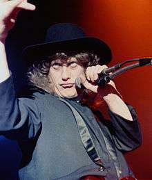 A man with curly hair holds a microphone to his face. He wears a black hat and jacket.