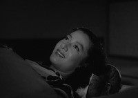 Noriko, in traditional costume, lying on a futon and covered with a blanket, stares at the ceiling, smiling