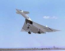White delta-wing aircraft taking off with landing gears retracting. At the front of aircraft are canards.