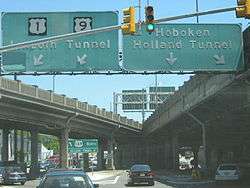 A ramp at a traffic light with a set of two overhead green signs. The one on the left reads U.S. Route 1/U.S, Route 9 north Lincoln Tunnel with two arrows pointing to the lower left and the one on the right reading Hoboken Holland Tunnel with a down arrow and an arrow pointing to the lower right.