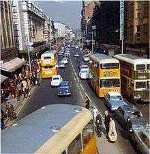 A busy urban street in Newcastle in the late 1960s. There are tall buildings on either side of the street, and various 1960s cars and buses on the road.