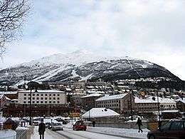 View of a town with buildings and streets covered with snow. In the background, behind the town, there are high, snowy mountains.