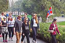 At right, an Asian woman holding a Norwegian flag with some writing on the bottom leads a group of casually dressed younger men and women, many wearing aviator sunglasses