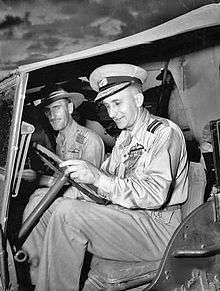 Two men in light-coloured military uniforms sitting in a jeep