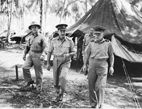 Three men in light-coloured military uniforms walking from tent, with palm trees in background