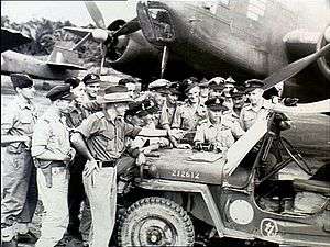 Approximately 15 men wearing military uniforms in discussion around a jeep, parked in front of a twin-engined aircraft