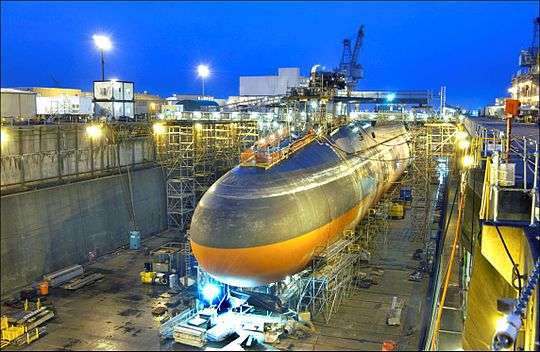 Photograph of the strategic missile submarine USS Ohio in drydock at the Puget Sound Naval Shipyard as night falls. The ship is surrounded by scaffolding and equipment, and several shipyard buildings appear in the background.