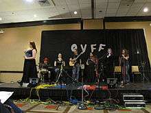 Photograph of several people on stage in front of a black curtain bearing the letters O V F F.