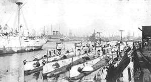 Nine submarines sit moored next to each other against a dock, with ships in the background