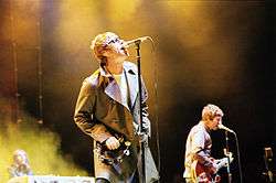 Liam Gallagher wearing sunglasses plays a tambourine and sings into a microphone. In the background, Noel Gallagher and Gem Archer play guitars and a keyboard respectively.