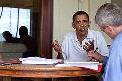 Obama, wearing a white shirt, is moving his hands while talking to a man in a blue shirt, who sits across him.