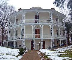 A three-story pale yellow house seen looking up its front walk on an overcast day with patchy snow in front. It has ornate white balconies on the front, where three faces are visible, and wooden doors.
