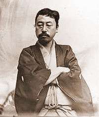 Portrait of an Asian man with moustache dressed in traditional Japanese cloths. He is looking down with his arms crossed.