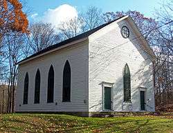 A white wooden building with a pointed roof and narrow green windows that end in pointed arches. Below one window in the front is a sign that says "Old Clove Church, 1787"