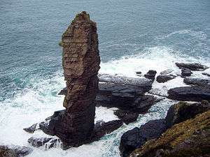 A tall sea stack consisting of a layered sedimentary rock  is situated off a rocky coast and sits amid breaking waves and foamy waters.