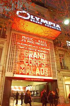 A photograph of the exterior of a theatre at night. Uppercase white text on the marquee reads "Olympia Bruno Coquatrix" against an orange backdrop. Several tree branches and leaves are visible at the top of the photograph on either side. A crowd of people are visible at the front entrance of the theatre.