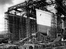 Photograph of a huge gantry with two openings in it, one of which is filled with the bows of a large ship that has been painted in light and dark colours.