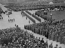 Black and white photo: Crowds of people and a ranked military guard gather to watch four men in ceremonial uniform approach a set of stone steps.