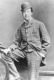 Oscar Wilde posing for a photograph, looking at the camera. He is wearing a checked suit and a bowler hat. His right foot is resting on a knee high bench, and his right hand, holding gloves, is on it. The left hand is in the pocket.