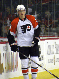 An ice hockey player is stood up straight by the edge of the rink. He is wearing a white uniform with orange shoulders and a stylised 'P' on his chest.
