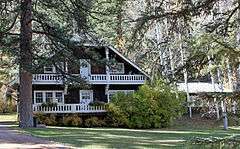 A dark brown two-story wooden house with white wooden trim and a pointed roof amid some tall trees.