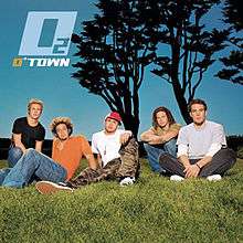 The band members are sitting on a grass field with a black-colored tree behind them in the blue sky background.